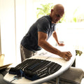Kelly Slater preparando su tabla Slater Designs en interior, con el grip trasero 5 Piece Action Pad negro y azul ya colocado en la cola, limpiando la superficie antes de pegar las piezas superiores.