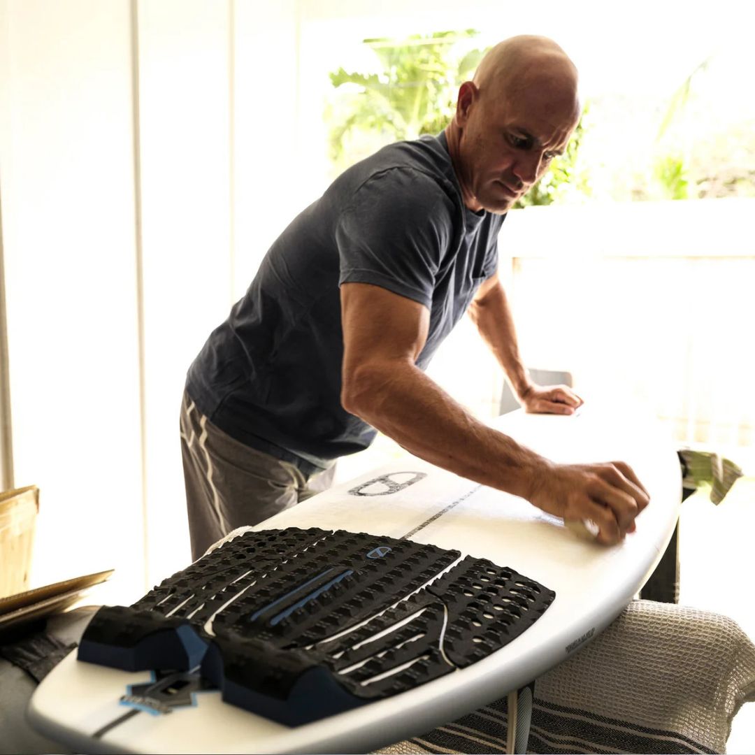 Kelly Slater preparando su tabla Slater Designs en interior, con el grip trasero 5 Piece Action Pad negro y azul ya colocado en la cola, limpiando la superficie antes de pegar las piezas superiores.