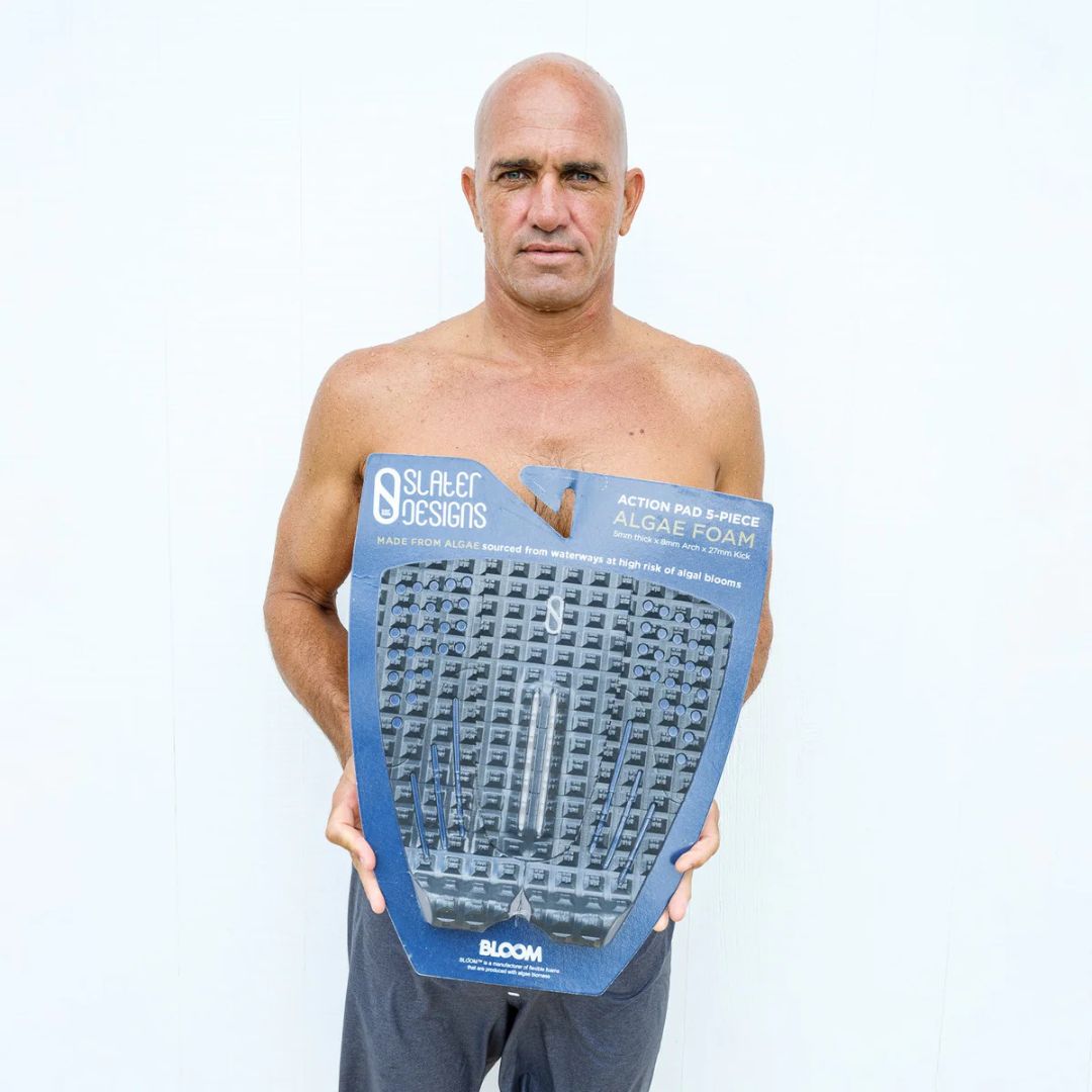 Kelly Slater sosteniendo el embalaje original del Slater Designs Action Pad 5 Piece Algae Foam con logo BLOOM azul, mostrando especificaciones 5mm grosor, arco 8mm y kick 27mm.