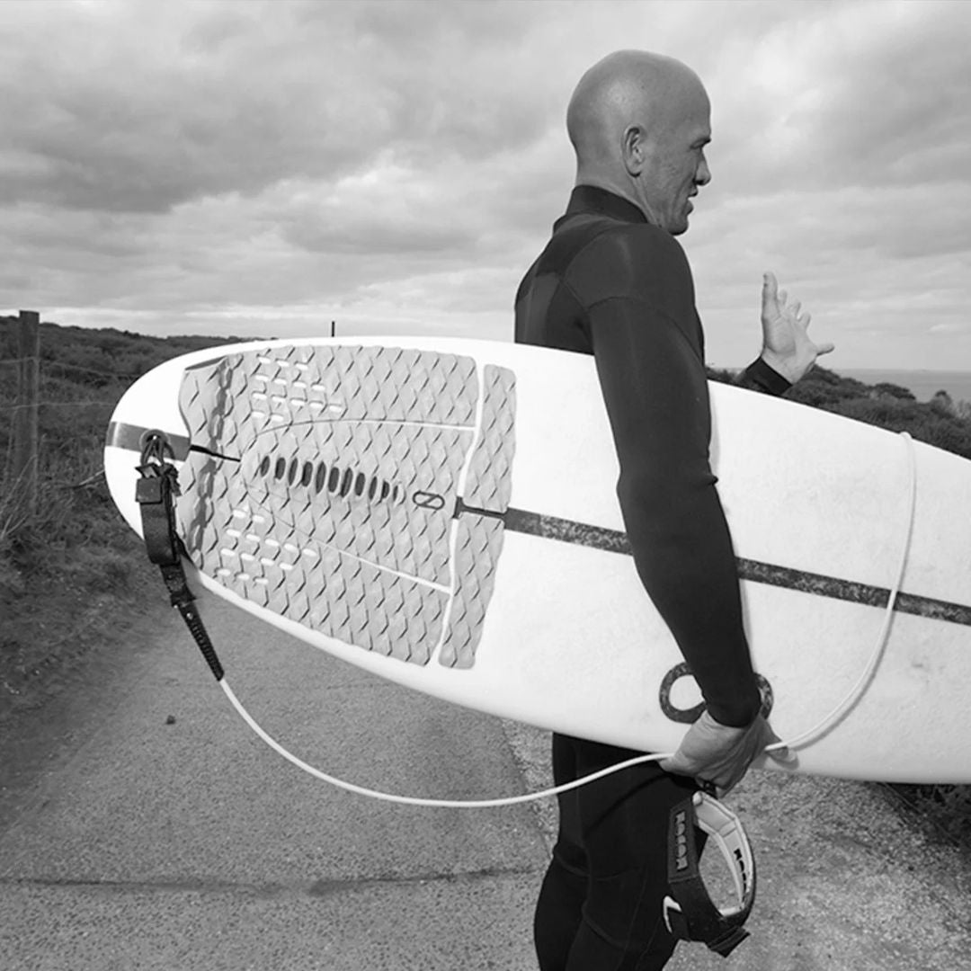 Kelly Slater de perfil en blanco y negro cargando tabla de surf con grip trasero Slater Designs 5 piezas visible, textura de diamante y construcción modular apreciable, camino costero al fondo.
