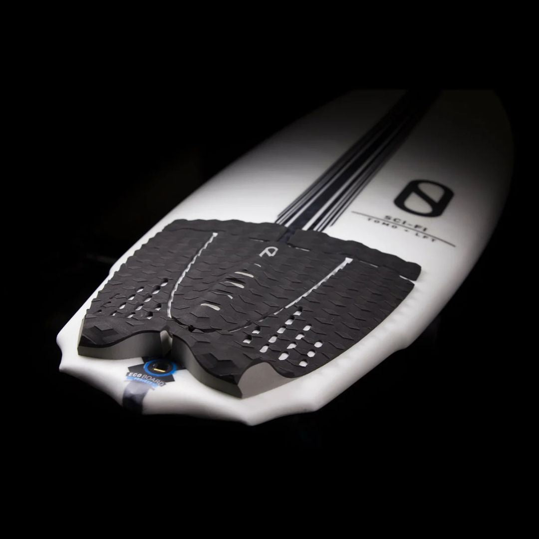Grip trasero Slater Designs 5 piezas negro en perspectiva sobre tabla Sci-Fi Tomo LFT blanca, fondo negro, mostrando cobertura total de cola, kick tail elevado y logo Slater Designs en el arco.