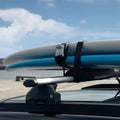 Dos tablas de SUP aseguradas con la cincha-candado Surflogic sobre las barras de techo del coche, con el mar y un muelle al fondo, hebilla bloqueable visible en el centro.