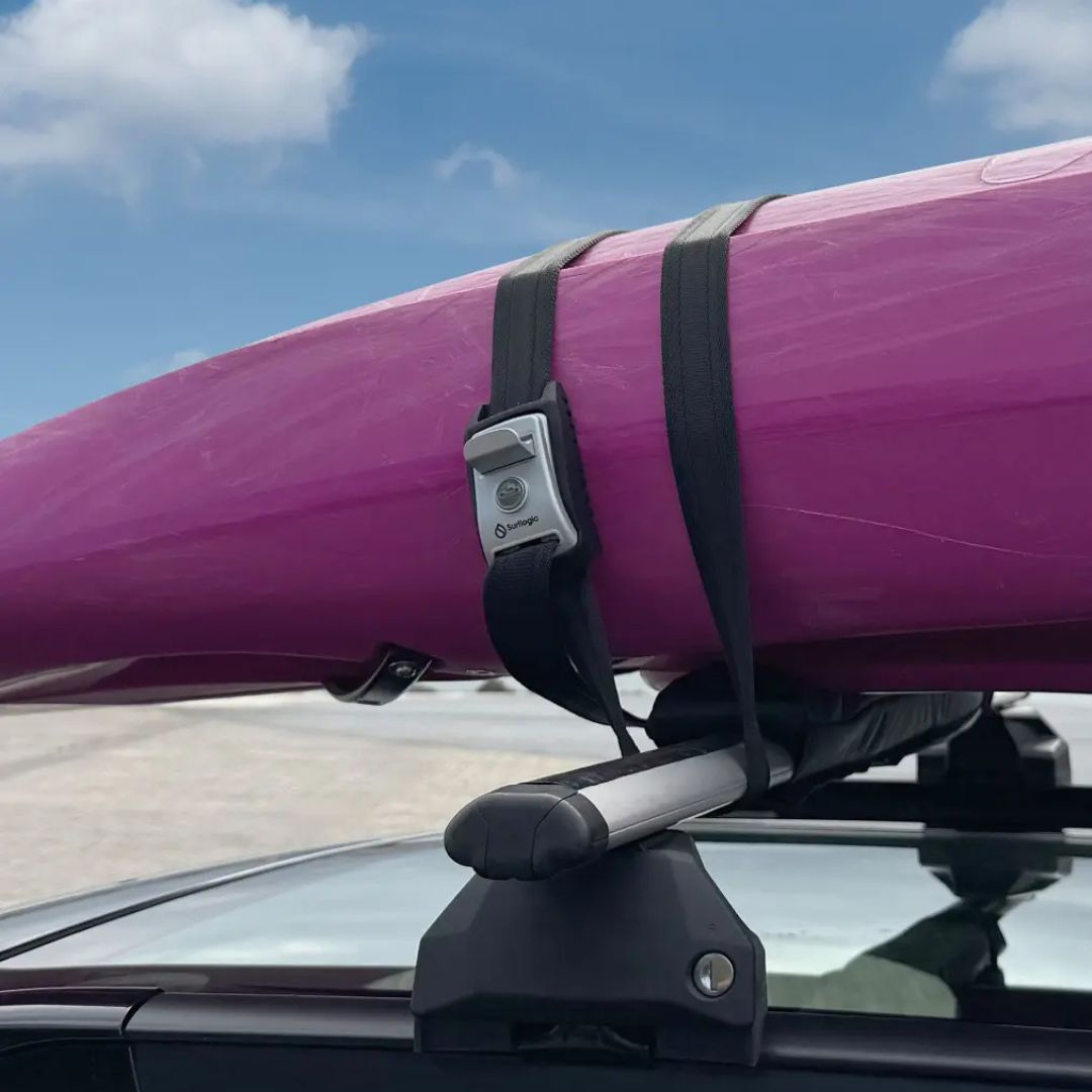 Cincha-candado Surflogic sujetando un kayak de color morado sobre la baca del techo del coche, con la hebilla metálica bloqueada visible en primer plano y cielo azul de fondo.