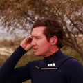 Surfista con traje de neopreno colocándose los tapones oídos surf Creatures SurfEars 4.0 black sage con cordón para prevenir el oído de surfista.
