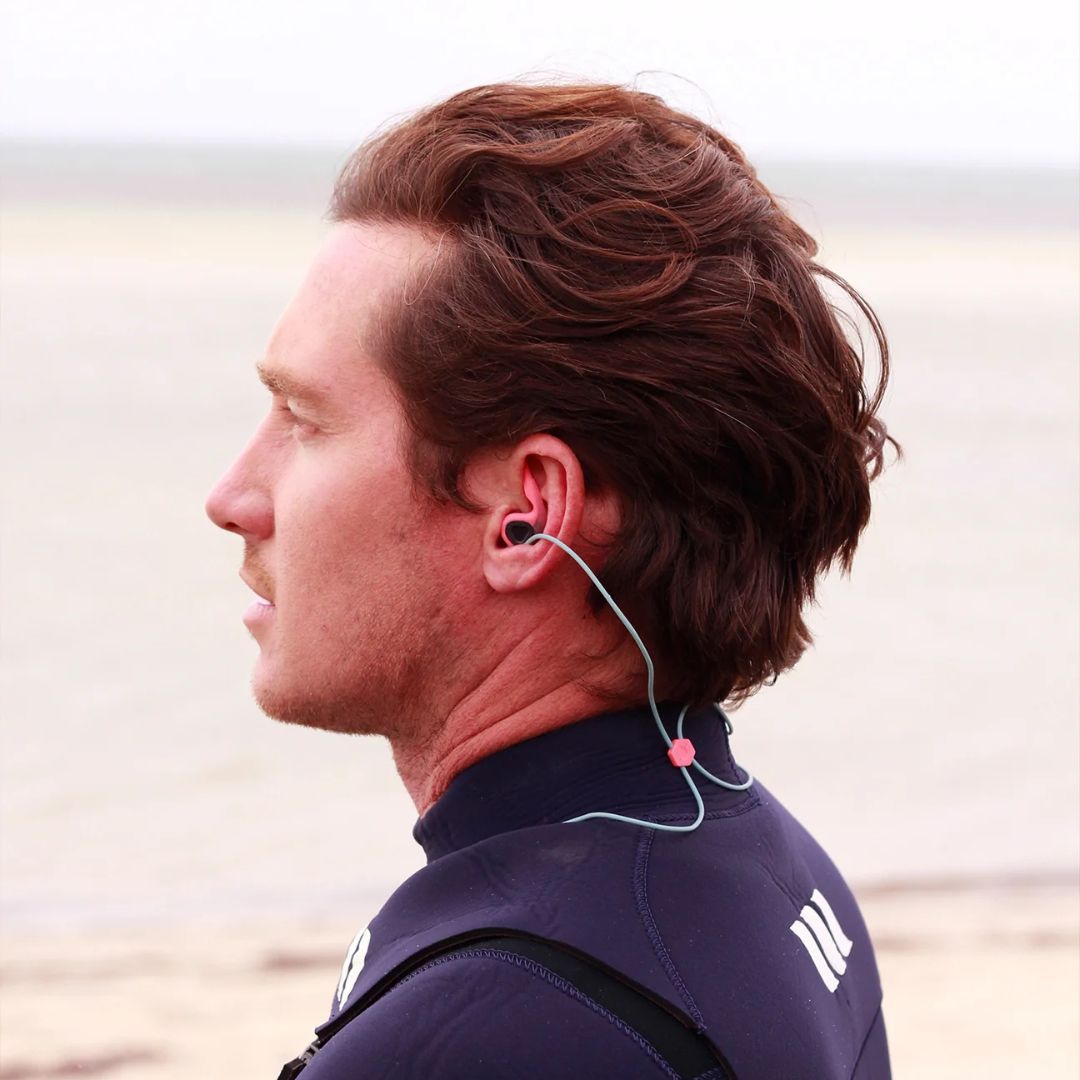 Surfista con traje de neopreno usando los tapones oídos surf SurfEars 4.0 coral sky con cordón sky azul para prevenir el oído de surfista en el agua.