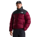 Plano medio frontal del modelo con la chaqueta de plumas The North Face Nuptse granate cerrada, cuello alto negro, logo bordado, bolsillos con cremallera y gorra negra.