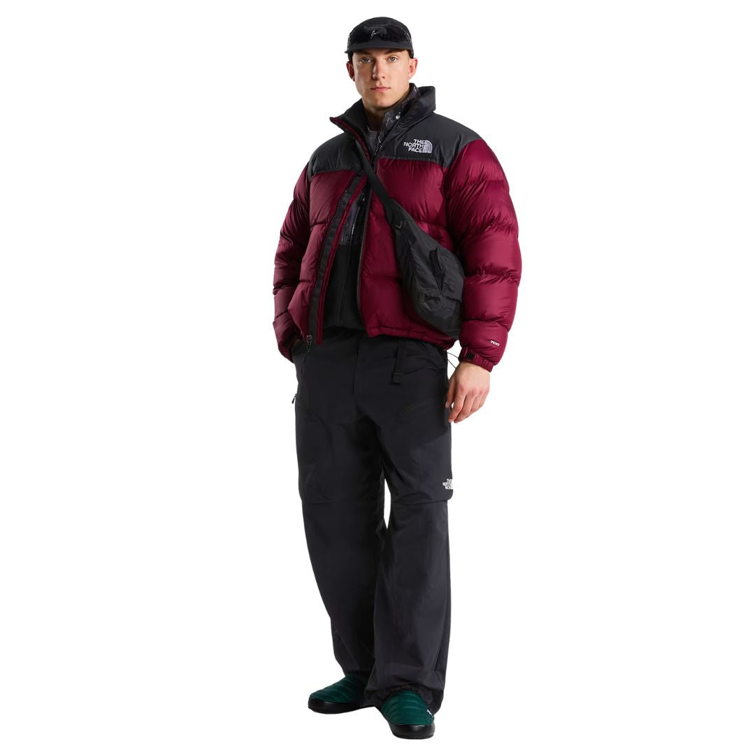 Modelo de cuerpo entero con la chaqueta de plumas The North Face Nuptse granate y negra, pantalón técnico oscuro, bandolera negra cruzada y zapatillas verdes.