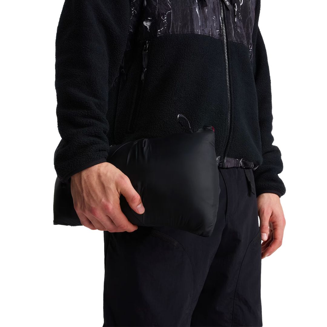 Modelo sosteniendo la chaqueta de plumas The North Face Nuptse plegada en su bolsa negra de transporte, con forro polar negro estampado y pantalón técnico oscuro.