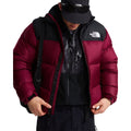 Modelo abriendo la cremallera de la chaqueta de plumas The North Face Nuptse granate sobre un forro polar negro estampado, con gorra negra, gafas y mochila al hombro.