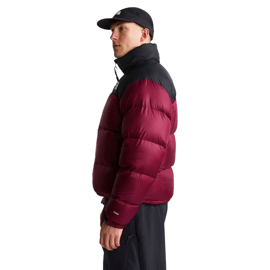 Perfil del modelo con la chaqueta de plumas The North Face Nuptse granate, de cuello alto negro, mangas acolchadas y número 700 en el puño, con gorra y pantalón negro.