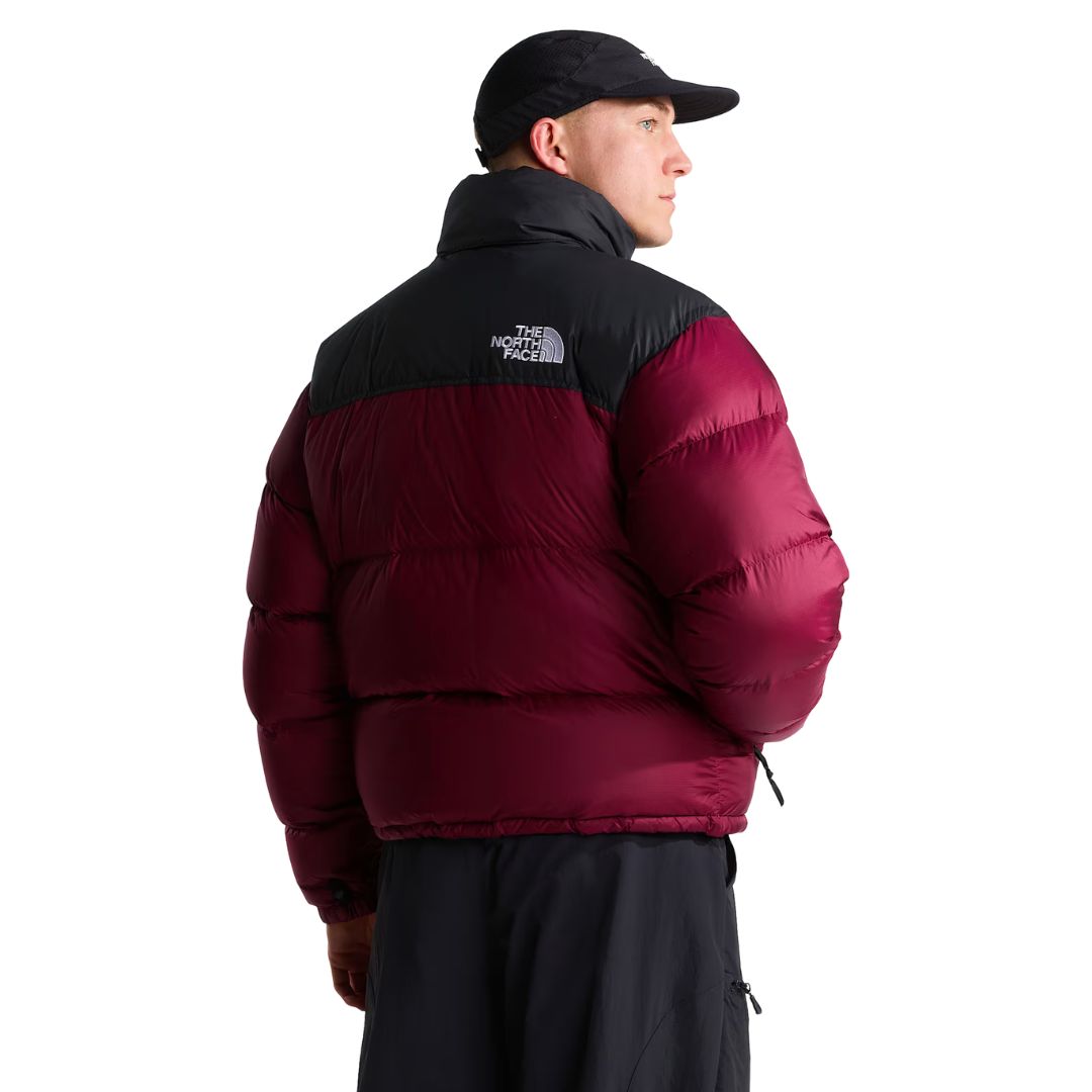 Espalda de la chaqueta de plumas The North Face Nuptse en granate con canesú negro y logo bordado, cuello alto y gorra negra; el modelo mira hacia un lado.