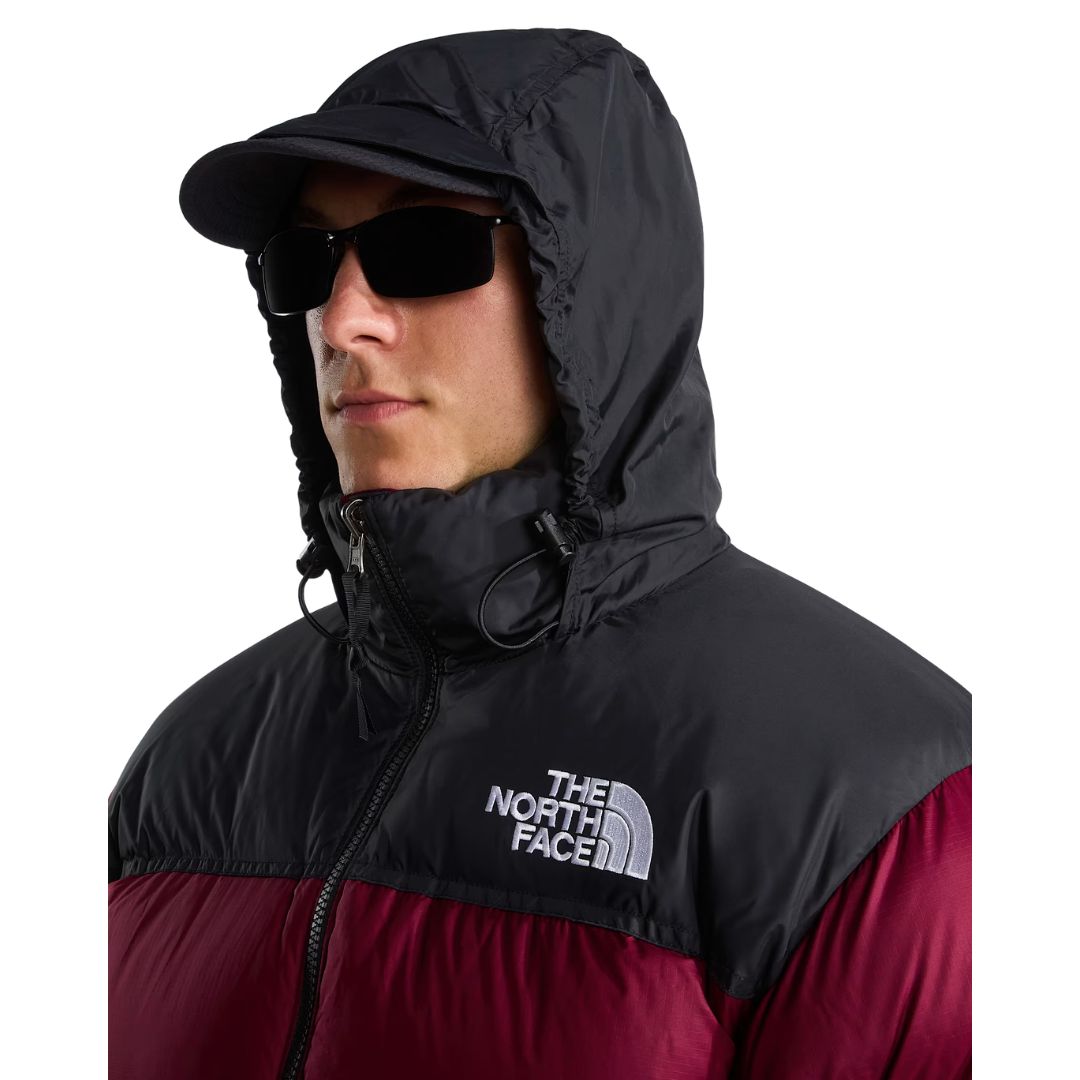 Primer plano de la capucha negra de la chaqueta de plumas The North Face Nuptse granate, con cordones de ajuste, logo bordado en el pecho, gorra y gafas de sol oscuras.