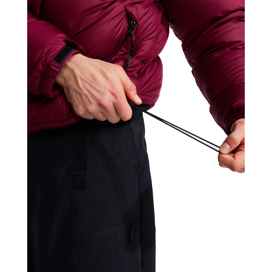 Detalle del cordón ajustable en el bajo de la chaqueta de plumas The North Face Nuptse granate, tirado con la mano sobre un pantalón técnico negro con cremalleras.