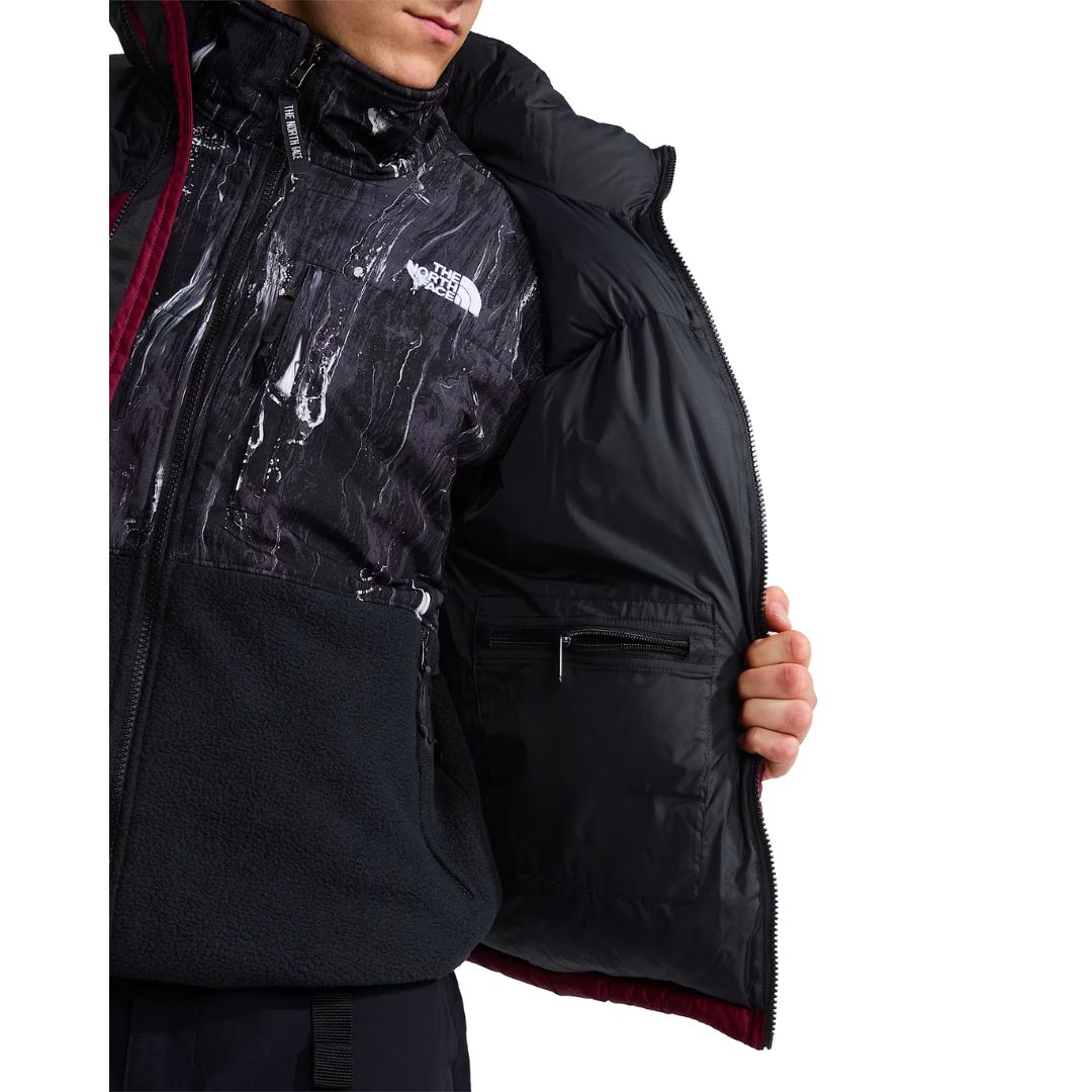Interior de la chaqueta de plumas The North Face Nuptse en granate, abierta para mostrar el bolsillo interior con cremallera y la capa de forro polar estampado y negro debajo.