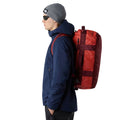 Hombre con chaqueta azul sosteniendo en la mano la bolsa The North Face Base Camp Voyager Duffel 42L color Mars Dust.