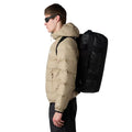 Hombre con chaqueta beige de perfil llevando en la espalda la bolsa The North Face Base Camp Voyager Duffel 42L en color TNF Black/Asphalt Grey.