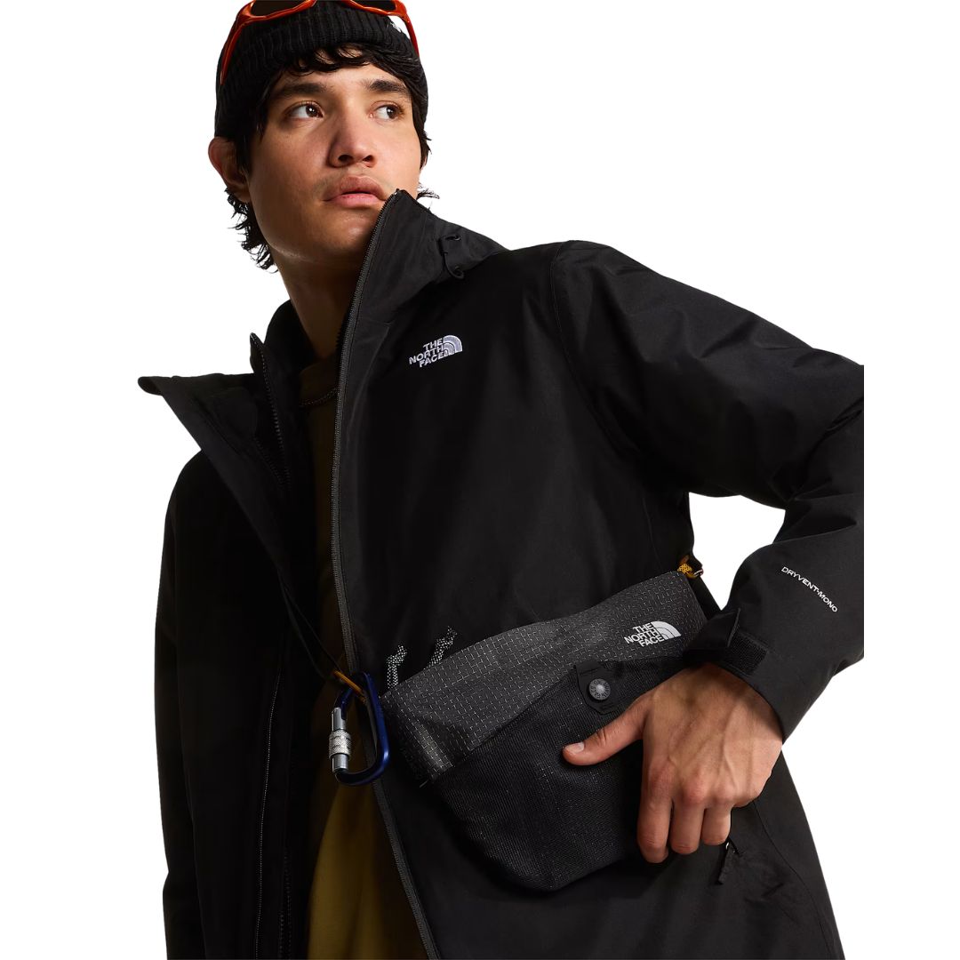 Modelo mirando de lado con la chaqueta The North Face Carto Mono Triclimate negra, bolso bandolera con mosquetón, gafas sobre el gorro y rotulado DRYVENT-MONO en la manga.