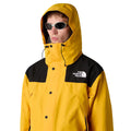Modelo vistiendo chaqueta The North Face Dryvent Mono Mountain Summit Gold y negro TNF con capucha puesta y gafas de sol.