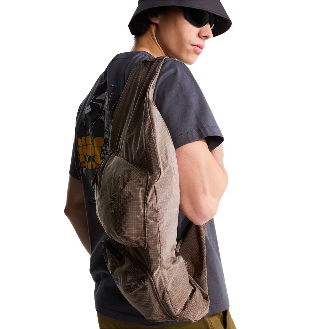 Camiseta The North Face Gear Up Get Out RLX en gris antracita puesta en modelo hombre con sombrero de pescador y gafas de sol, luciendo bandolera color topo cruzada al hombro sobre el gráfico trasero.