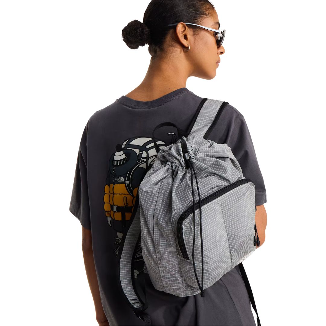 Camiseta The North Face Gear Up Get Out RLX en gris antracita puesta en modelo mujer, vista de espalda con mochila gris a cuadros de la marca al hombro, luciendo parcialmente el gráfico trasero.