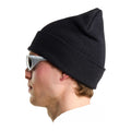 Perfil de un modelo con gafas de sol y sudadera clara que lleva puesto el gorro The North Face Red Box Beanie TNF Black, con el borde vuelto y la copa de punto vista de lado