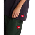 Camiseta The North Face Red Box oversize color obsidian, primer plano del bajo con el parche bordado rojo THE NORTH FACE junto a un pantalón verde y chaqueta al brazo.