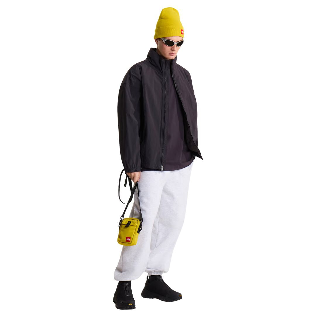Look completo con pantalón de chándal The North Face Red Box en gris jaspeado TFN Classic, combinado con chaqueta cortavientos negra, gorro amarillo y bandolera a juego.