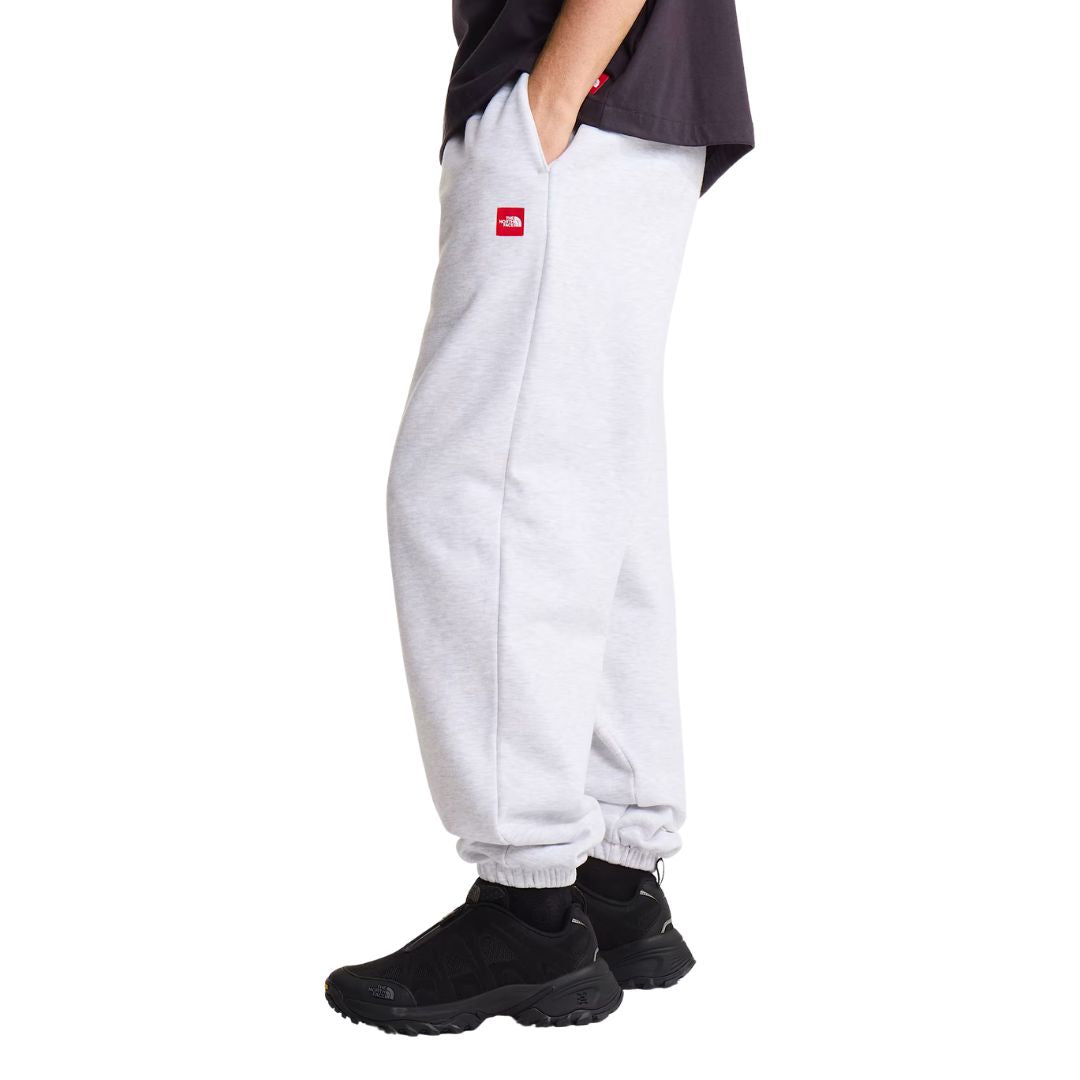 Pantalón de chándal The North Face Red Box en gris jaspeado TFN Classic, vista lateral de cuerpo completo con camiseta negra, mano en el bolsillo y parche rojo del logo en la cadera.