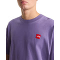 Detalle del parche bordado de la camiseta The North Face Redbox unisex color Grounded Plum, primer plano del cuello y el logo rojo en el pecho del hombre.
