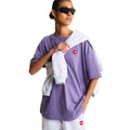 Camiseta The North Face Redbox unisex color Grounded Plum, mujer con gafas de sol sujetando una sudadera gris sobre el hombro, parche bordado rojo en el pecho.