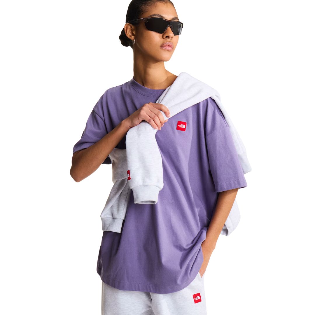 Camiseta The North Face Redbox unisex color Grounded Plum, mujer con gafas de sol sujetando una sudadera gris sobre el hombro, parche bordado rojo en el pecho.
