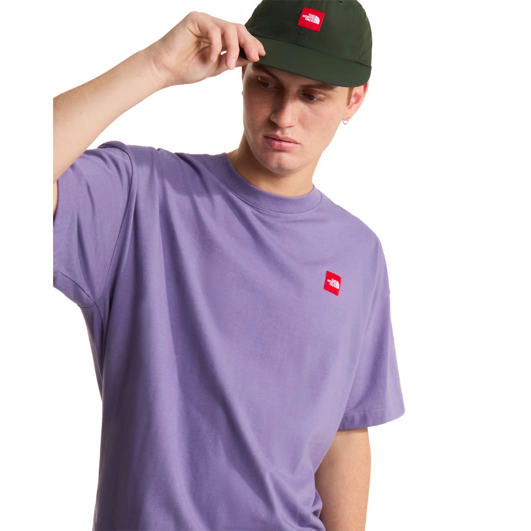 Camiseta The North Face Redbox manga corta unisex color Grounded Plum, hombre ajustándose una gorra verde, parche bordado rojo visible en el pecho izquierdo.