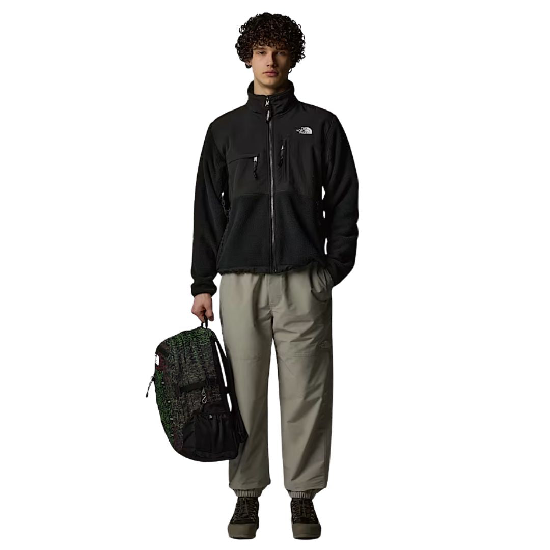 Hombre con la Chaqueta The North Face Retro Denali para hombre en color TFN Black con el logo en blanco, vista frontal completa con cremallera cerrada y mochila colgada