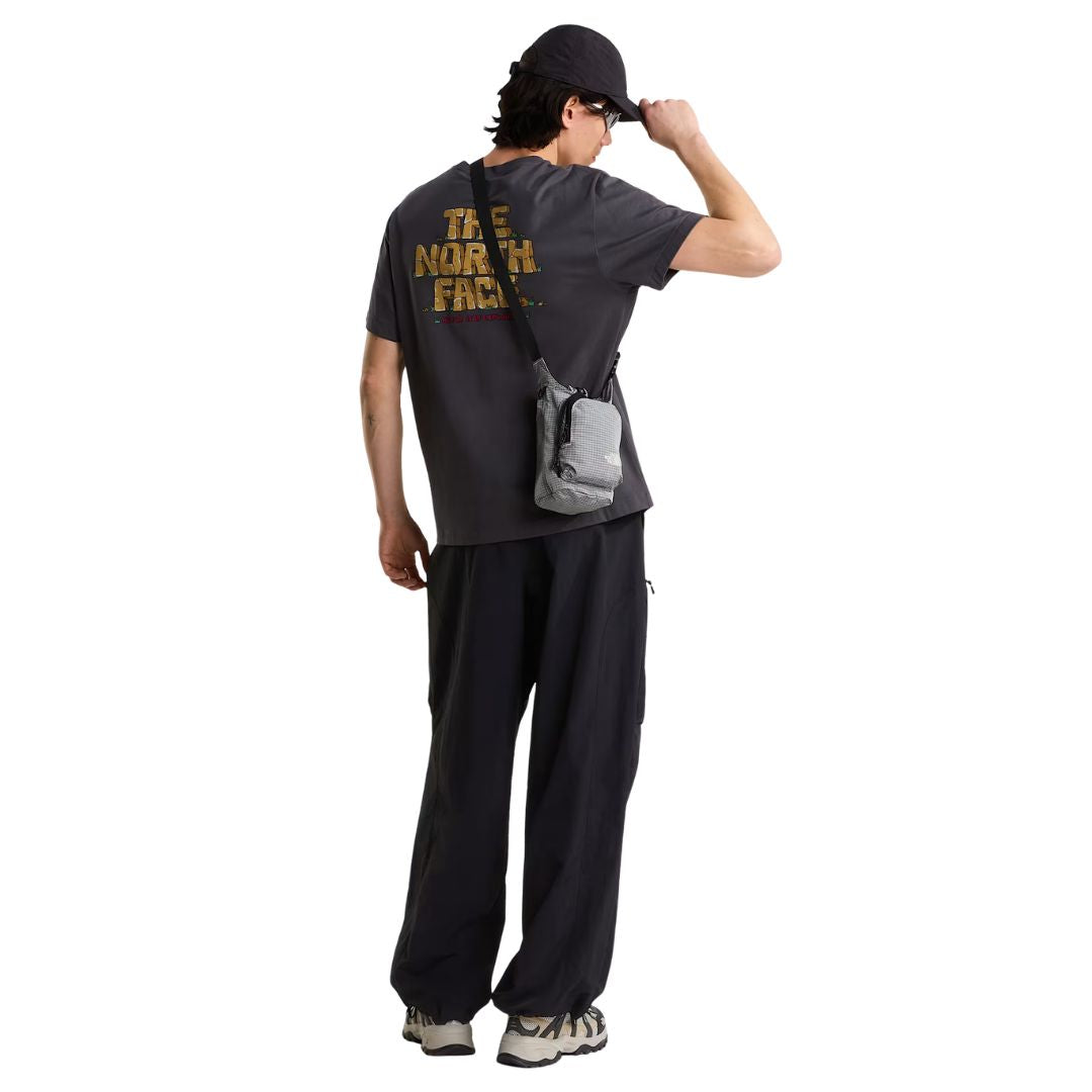 Camiseta The North Face Stone Logo RLX en gris antracita en modelo hombre de cuerpo entero por detrás, combinada con pantalón ancho negro y bandolera gris a juego, luciendo el estampado trasero.