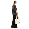 Camiseta The North Face Stone Logo RLX en gris antracita en modelo mujer de cuerpo entero por detrás, combinada con pantalón negro ancho y bolso tote beige de la marca.