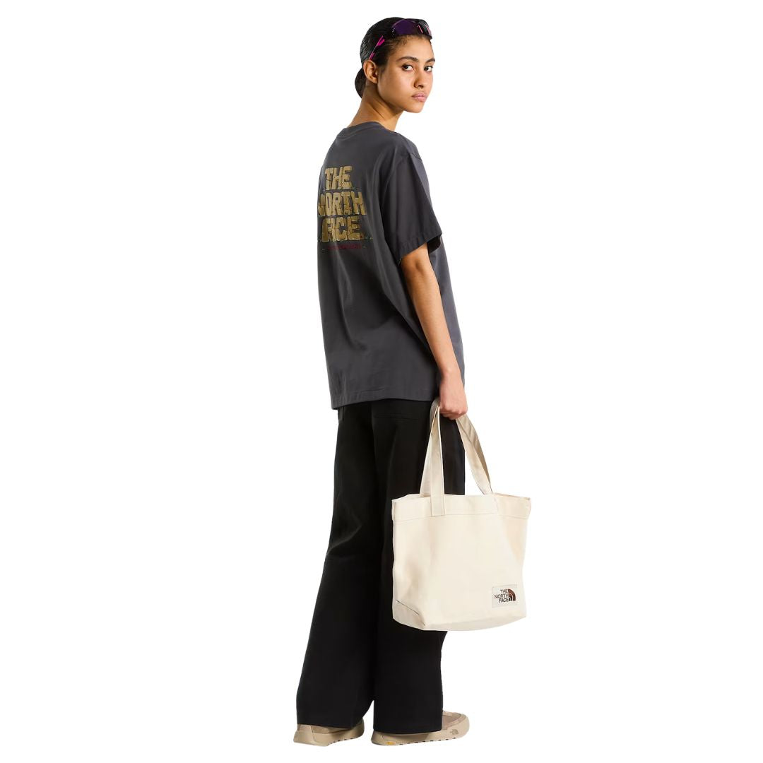 Camiseta The North Face Stone Logo RLX en gris antracita en modelo mujer de cuerpo entero por detrás, combinada con pantalón negro ancho y bolso tote beige de la marca.
