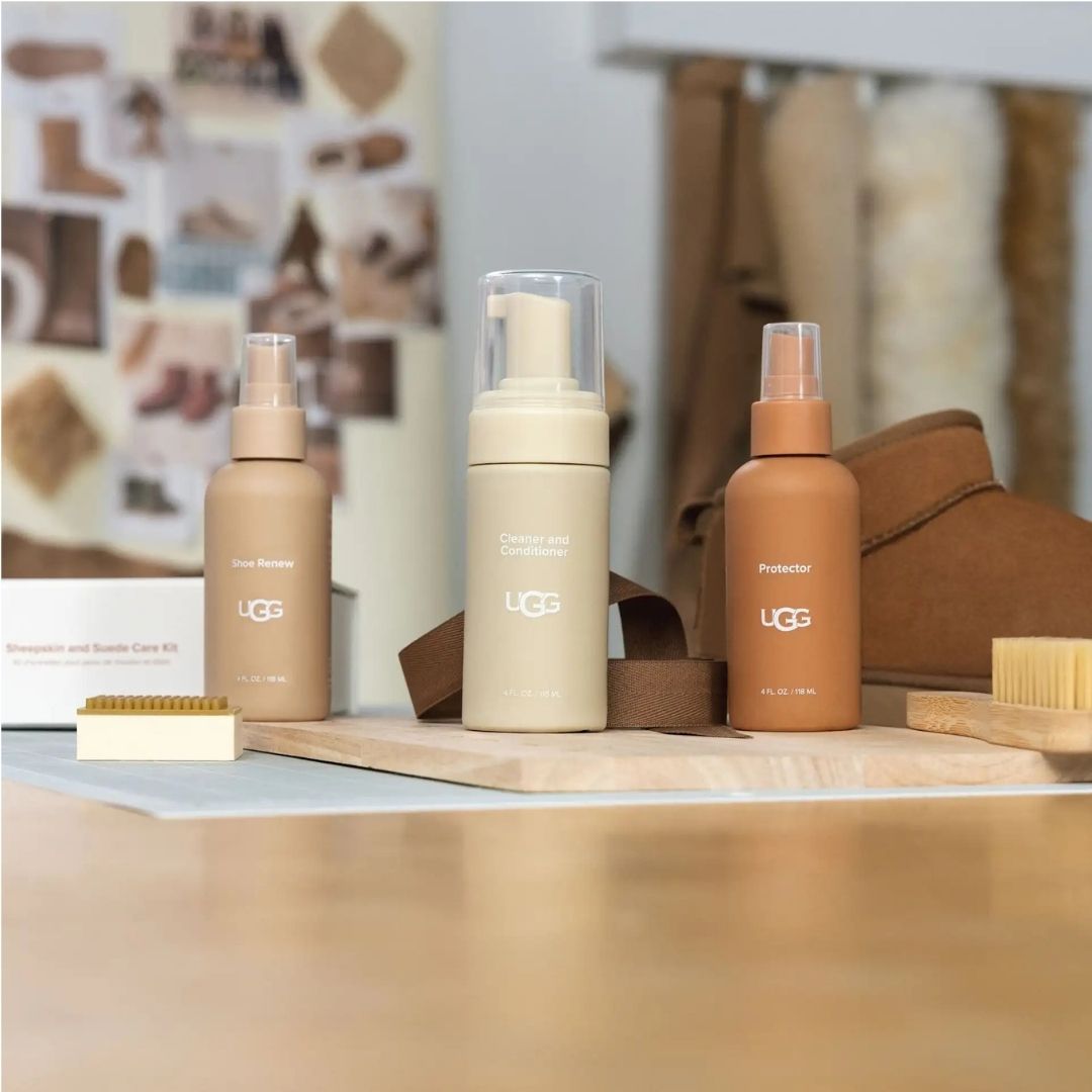 Botes del UGG Care Kit Cleaner Shoe Renew, Cleaner and Conditioner y Protector sobre una tabla de madera junto al cepillo de bambú, con calzado de piel de oveja al fondo.