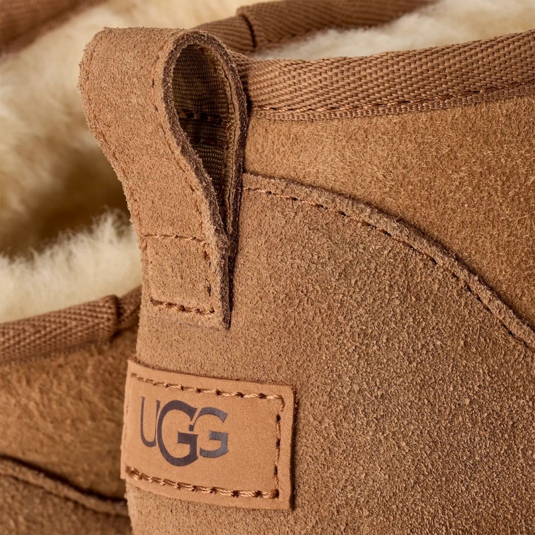 Primer plano del talón de la bota UGG Classic Ultra Mini en color chestnut con la etiqueta de piel grabada con el logotipo UGG y las costuras decorativas sobrehiladas.