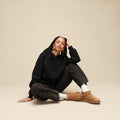 Mujer sentada en el suelo con botas UGG Classic Ultra Mini en color chestnut combinadas con sudadera negra y vaqueros gastados, mostrando la caña baja y la piel de oveja.