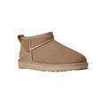 Vista en ángulo de tres cuartos de la bota UGG Classic Ultra Mini en color sand destacando la puntera redondeada, la textura aterciopelada y la suela EVA antideslizante.