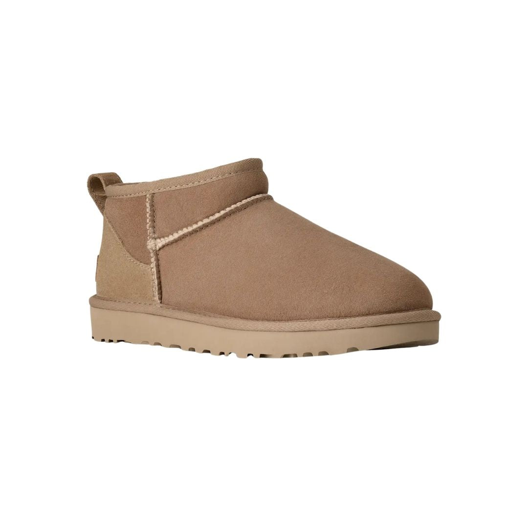Vista en ángulo de tres cuartos de la bota UGG Classic Ultra Mini en color sand destacando la puntera redondeada, la textura aterciopelada y la suela EVA antideslizante.