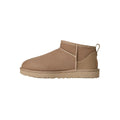 Vista lateral de la bota UGG Classic Ultra Mini en color sand mostrando la línea superior asimétrica, el pespunte central decorativo y la silueta de caña ultra corta.