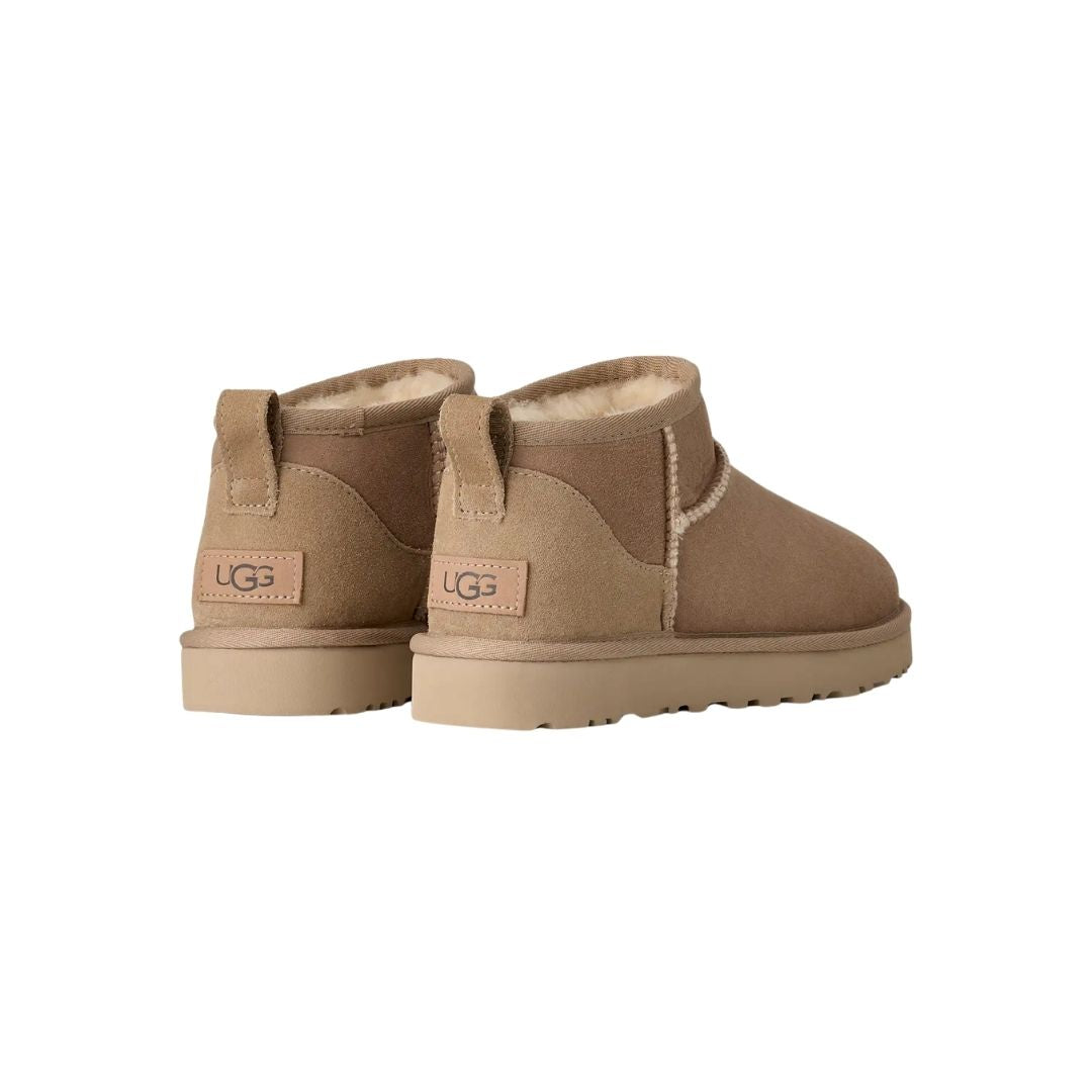 Par de botas UGG Classic Ultra Mini en color sand vistas desde atrás en ángulo, mostrando la trabilla trasera, el contrafuerte de ante y la suela flexible de goma.