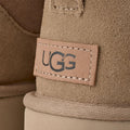 Primer plano del talón de la bota UGG Classic Ultra Mini en color sand con la etiqueta de piel grabada con el logotipo UGG y las costuras decorativas sobrehiladas.