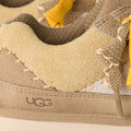 Detalle de costuras y logotipo en zapatillas UGG Lo Lowmel Biarritz color mustard seed con empeine de ante y cordones amarillos.