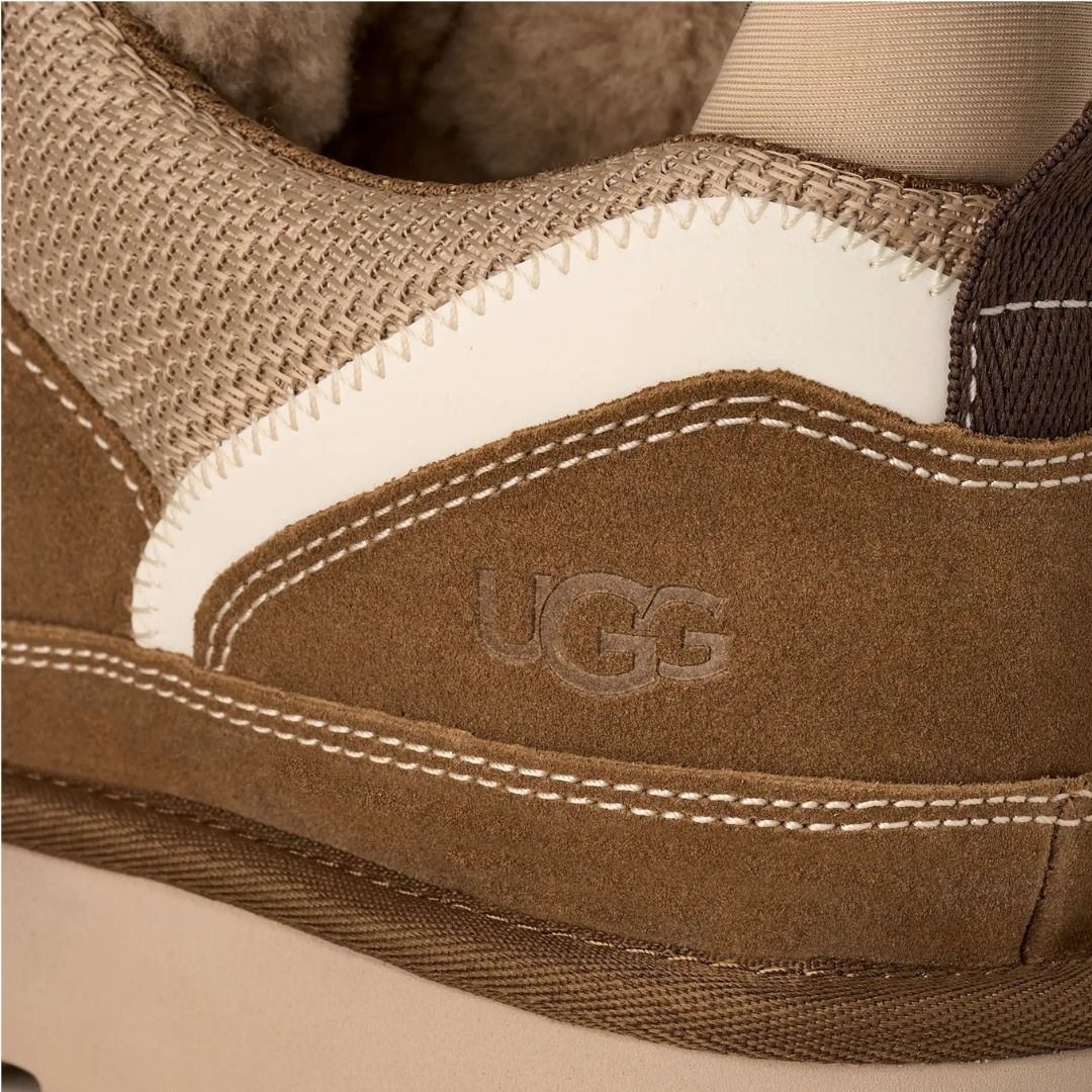 Primer plano del lateral de la zapatilla UGG Lowmel en color light bronze con el logotipo UGG grabado en ante y la combinación de malla técnica y textil crudo.