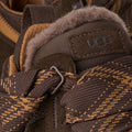 Primer plano del talón de la zapatilla UGG Lowmel en color dusted cocoa con el logotipo UGG grabado, la hebilla del cordón y el forro de piel visible.