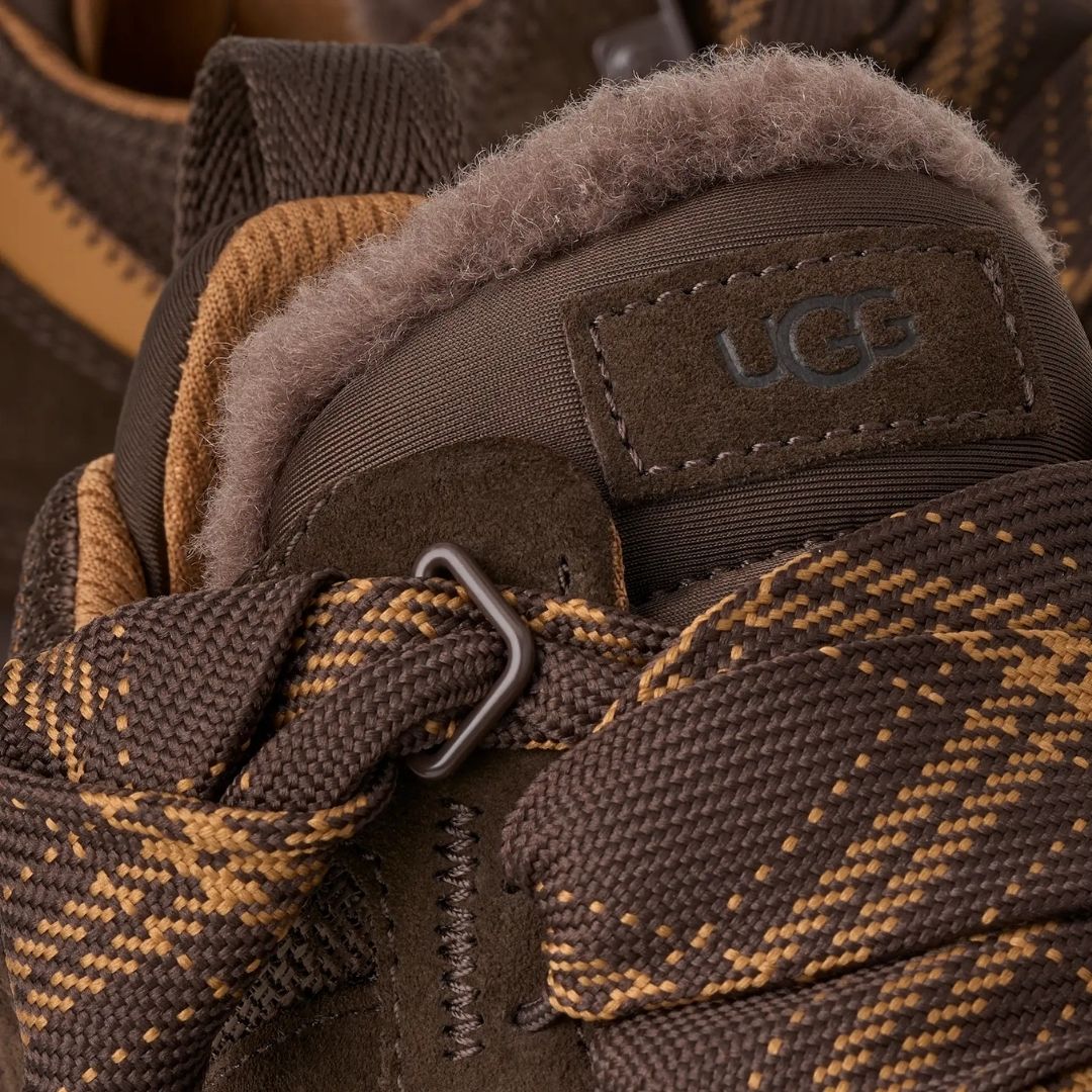 Primer plano del talón de la zapatilla UGG Lowmel en color dusted cocoa con el logotipo UGG grabado, la hebilla del cordón y el forro de piel visible.
