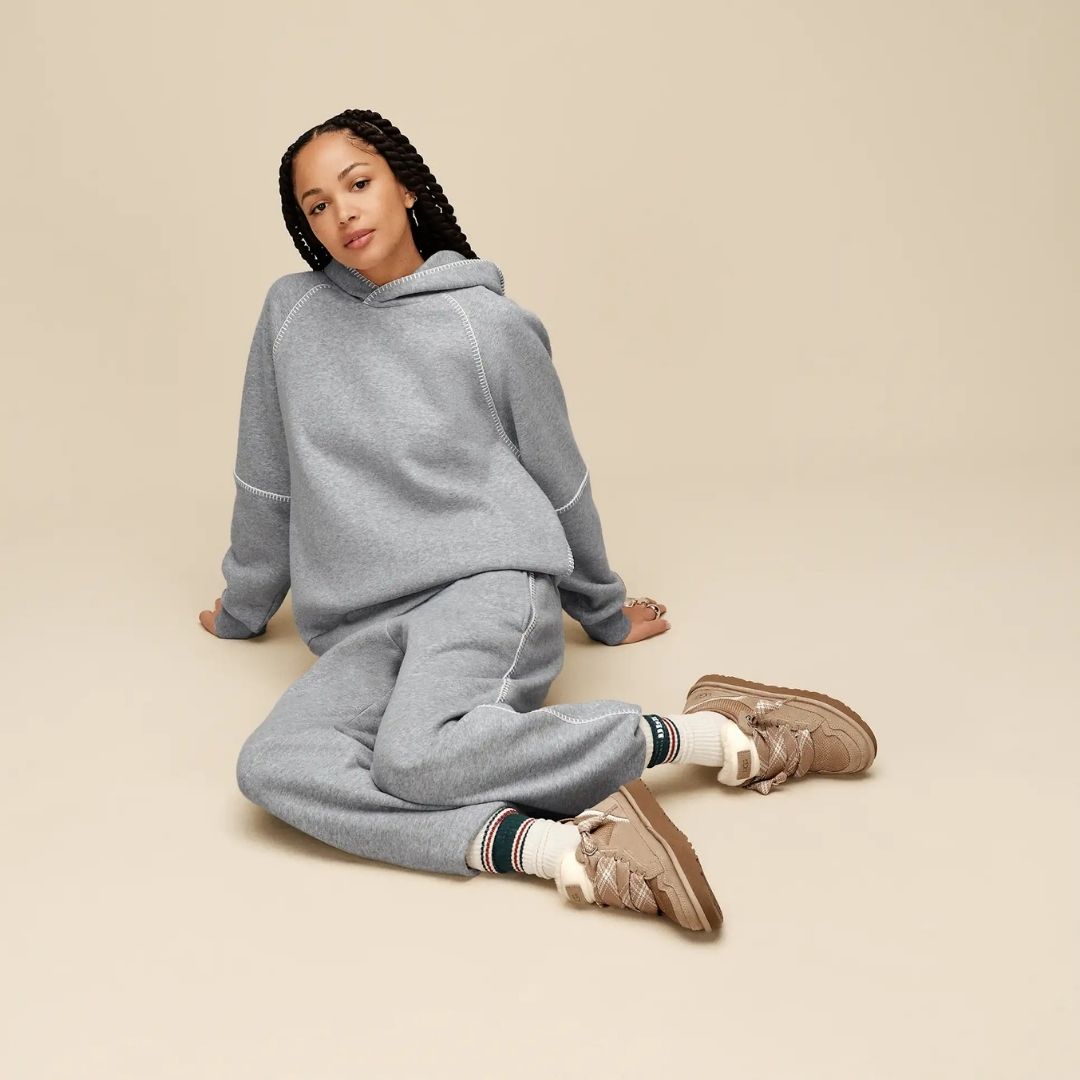 Mujer sentada con zapatillas UGG Lowmel en color sand combinadas con chándal gris, luciendo los cordones gruesos anudados y el empeine de ante y malla.