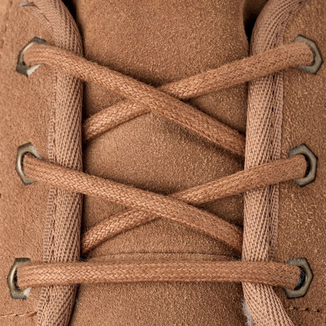 Primer plano de los cordones de algodón cruzados en los ojetes metálicos de la bota chukka UGG Neumel en color chestnut sobre el empeine de ante suave.